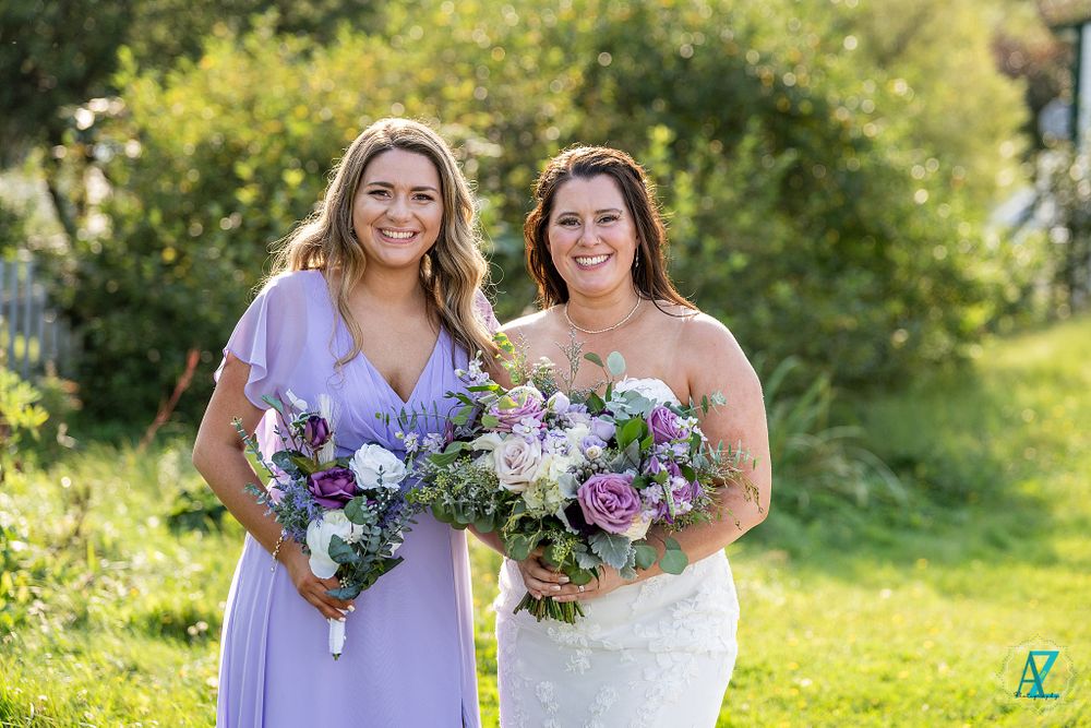 Wedding party members smile in lavender dress and white gown while holding coordinated floral bouquets.
