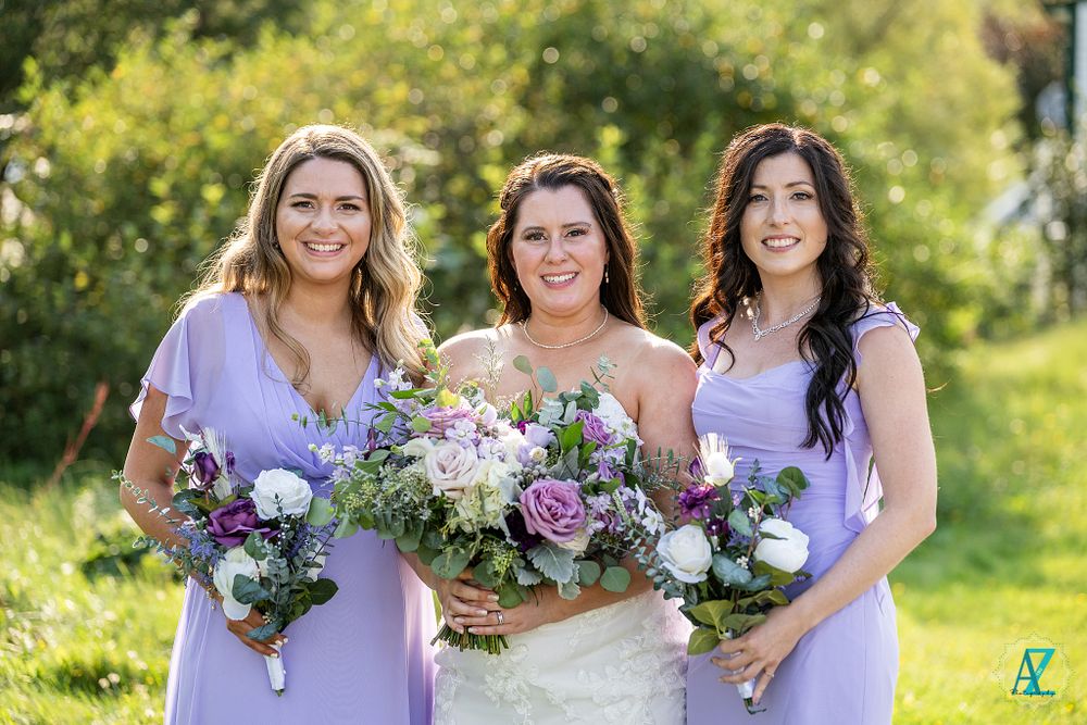 Three wedding attendants pose with coordinating purple and white bouquets in outdoor garden setting.