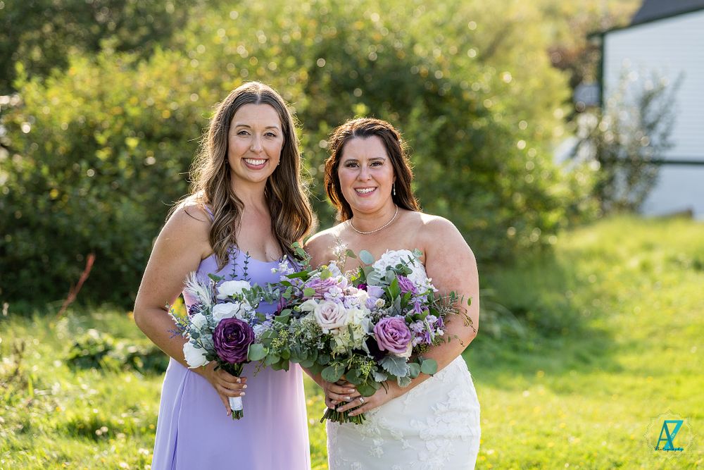 Two wedding attendants share a joyful moment while holding purple and white bouquets outdoors.
