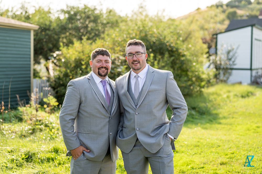 Two wedding party members in matching gray suits pose in sunny outdoor setting.
