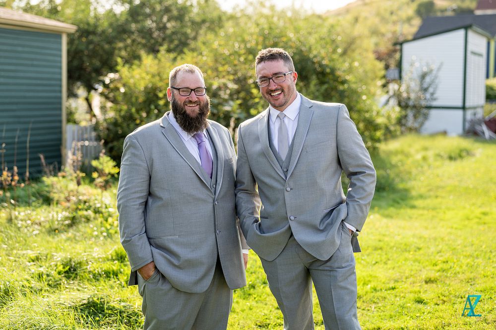 Two people in matching light gray suits pose on grass with green buildings in background.
