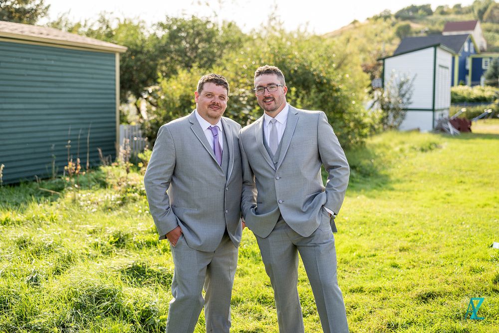 Two people in identical gray suits share a candid moment while standing on sunny lawn.