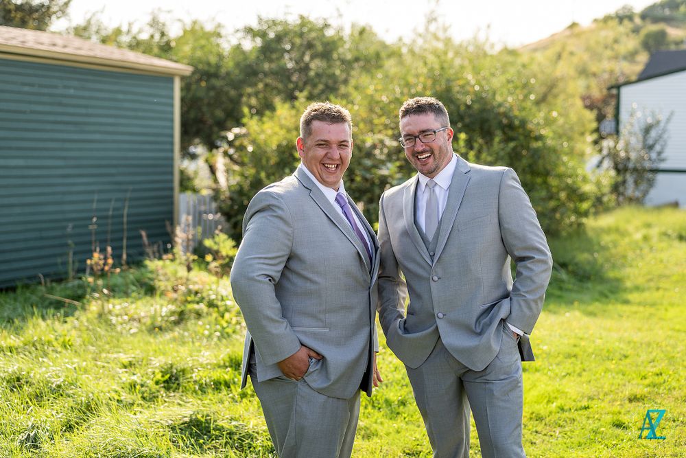 Two people in coordinating gray suits laugh together outdoors on a grassy hill.