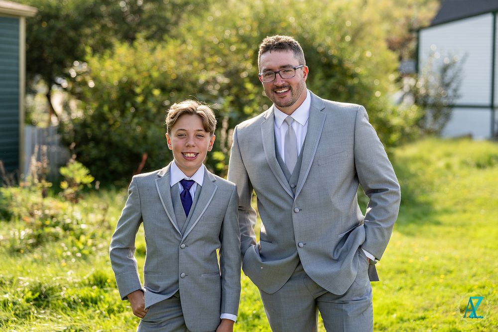 Two people in matching gray suits pose together on grass with buildings behind them.