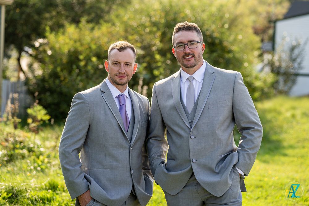 Two people wearing gray suits and purple accents pose outside on sunny grass.