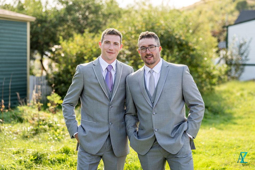 Two people in matching gray suits with purple accessories pose on sunny grass.