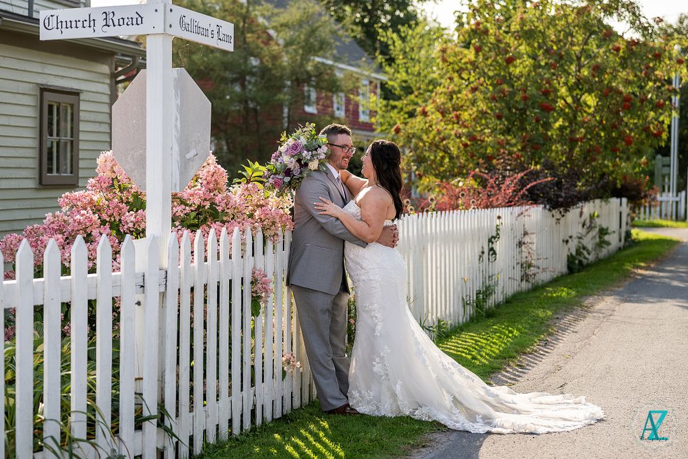A wedding couple stands by a white picket fence and signpost on a sunny garden path.