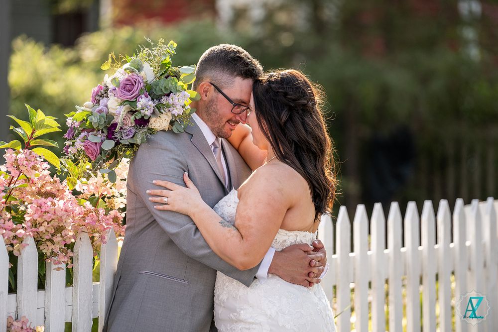 A couple embraces in front of white picket fence with colorful floral arrangements.