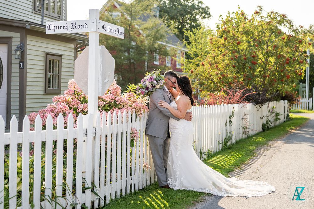 A wedding couple embraces near a white fence post surrounded by pink flowers and greenery.