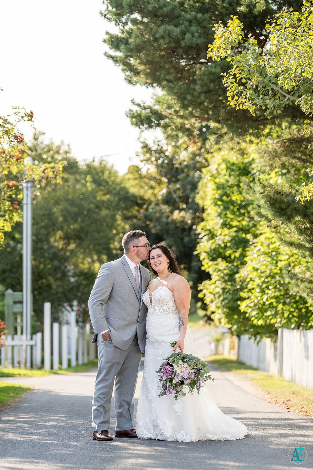 Newlyweds pose on a sunlit path with white picket fencing and lush summer foliage.