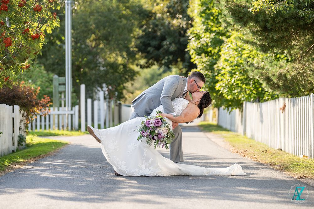 A romantic dip pose between newlyweds on a tree-lined path with white fencing.