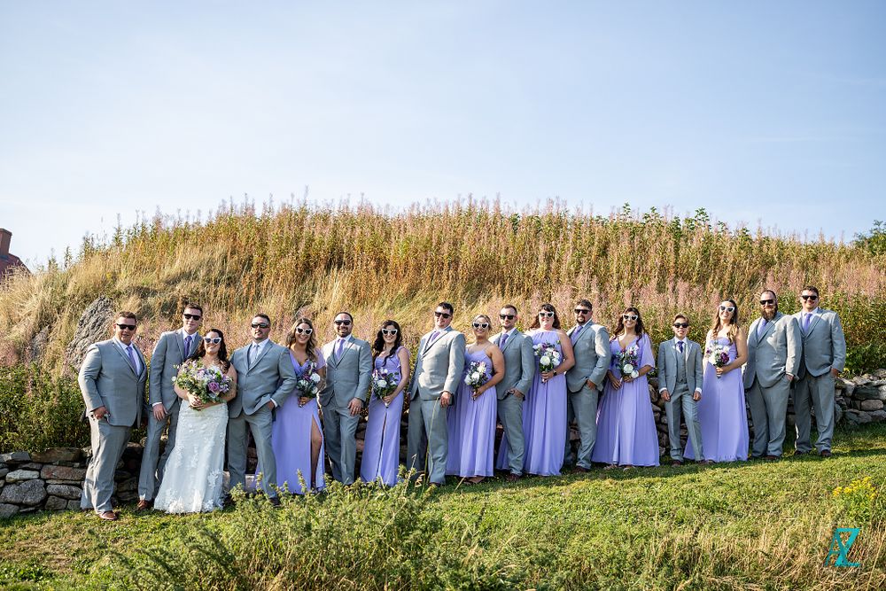 Large wedding party in purple and grey attire poses on a grassy hillside.