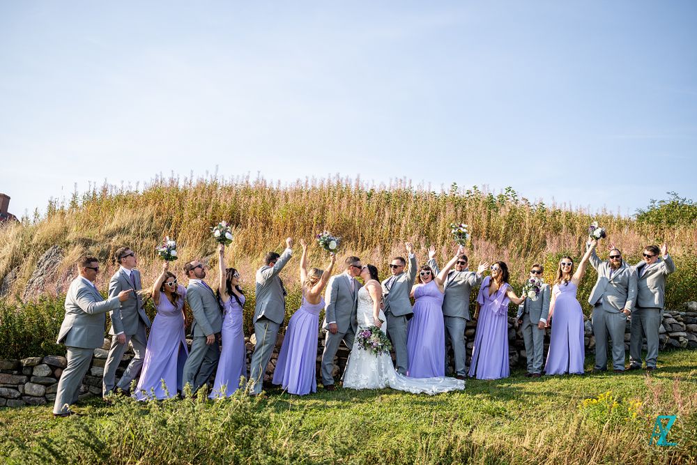 Wedding party celebrates with fun poses on a sunny hillside wearing purple and grey.