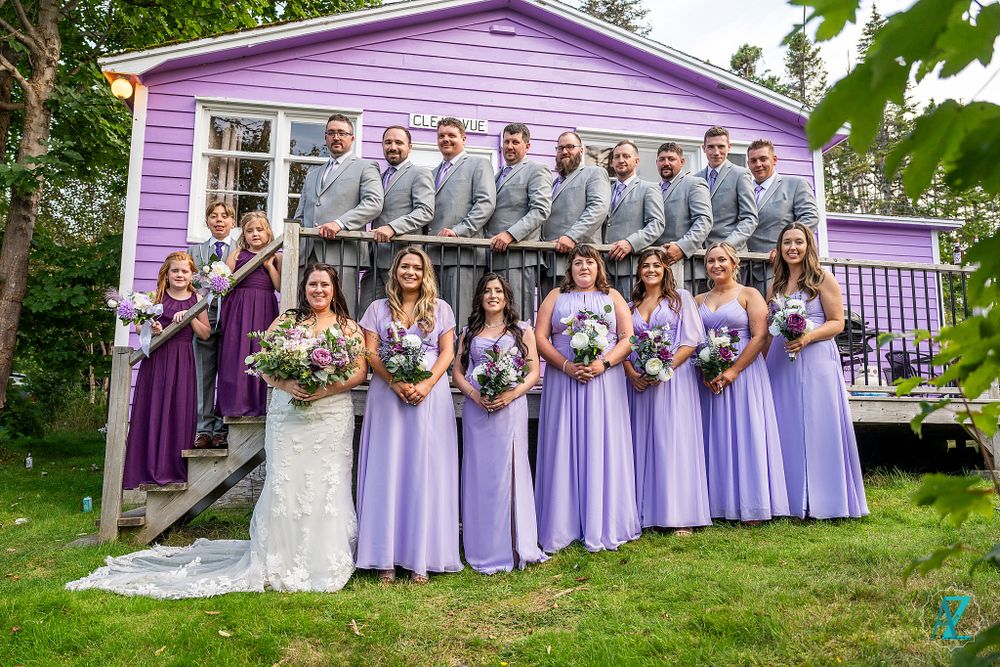 Wedding party in purple and grey attire poses in front of a charming lilac colored house.