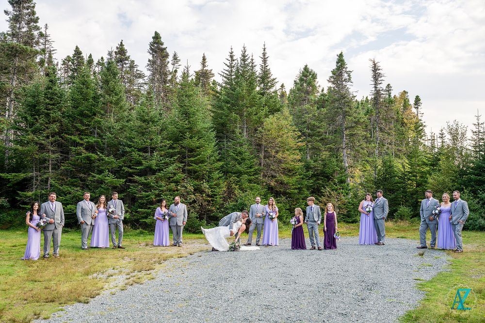 Wedding party in lavender dresses poses on a gravel path surrounded by tall evergreen trees.