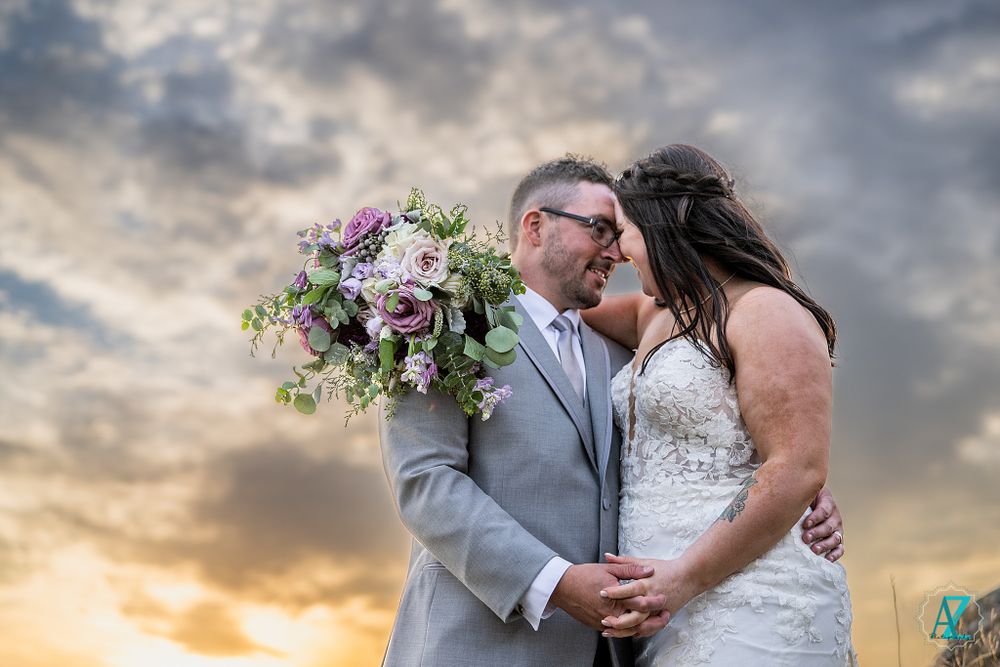 Dramatic sunset wedding portrait with purple and white bouquet against cloudy sky.