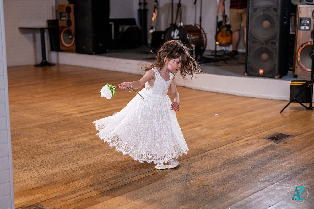 Young flower girl in white lace dress twirls on wooden dance floor at wedding reception.