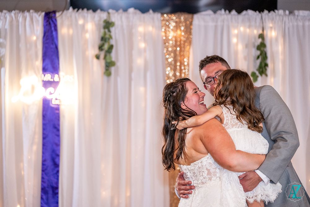 A romantic wedding couple sharing a kiss during their first dance beneath twinkling lights and purple decor.