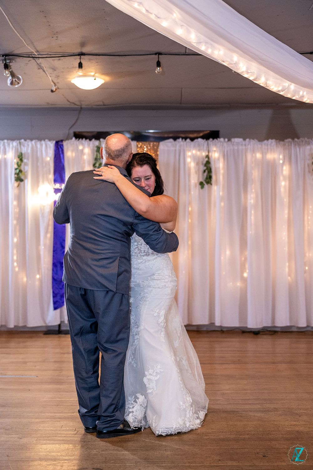 Intimate wedding dance moment captured against white curtain backdrop with string lights.