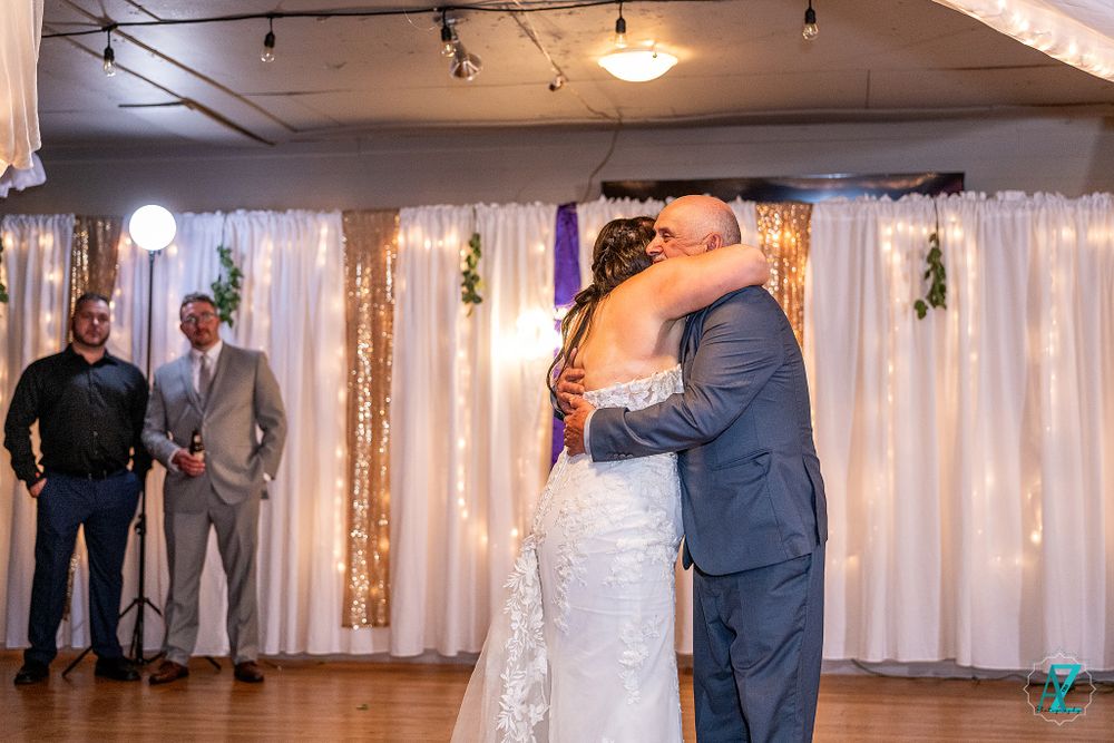 Elegant wedding couple embraces during first dance in front of white draped backdrop with twinkling lights.