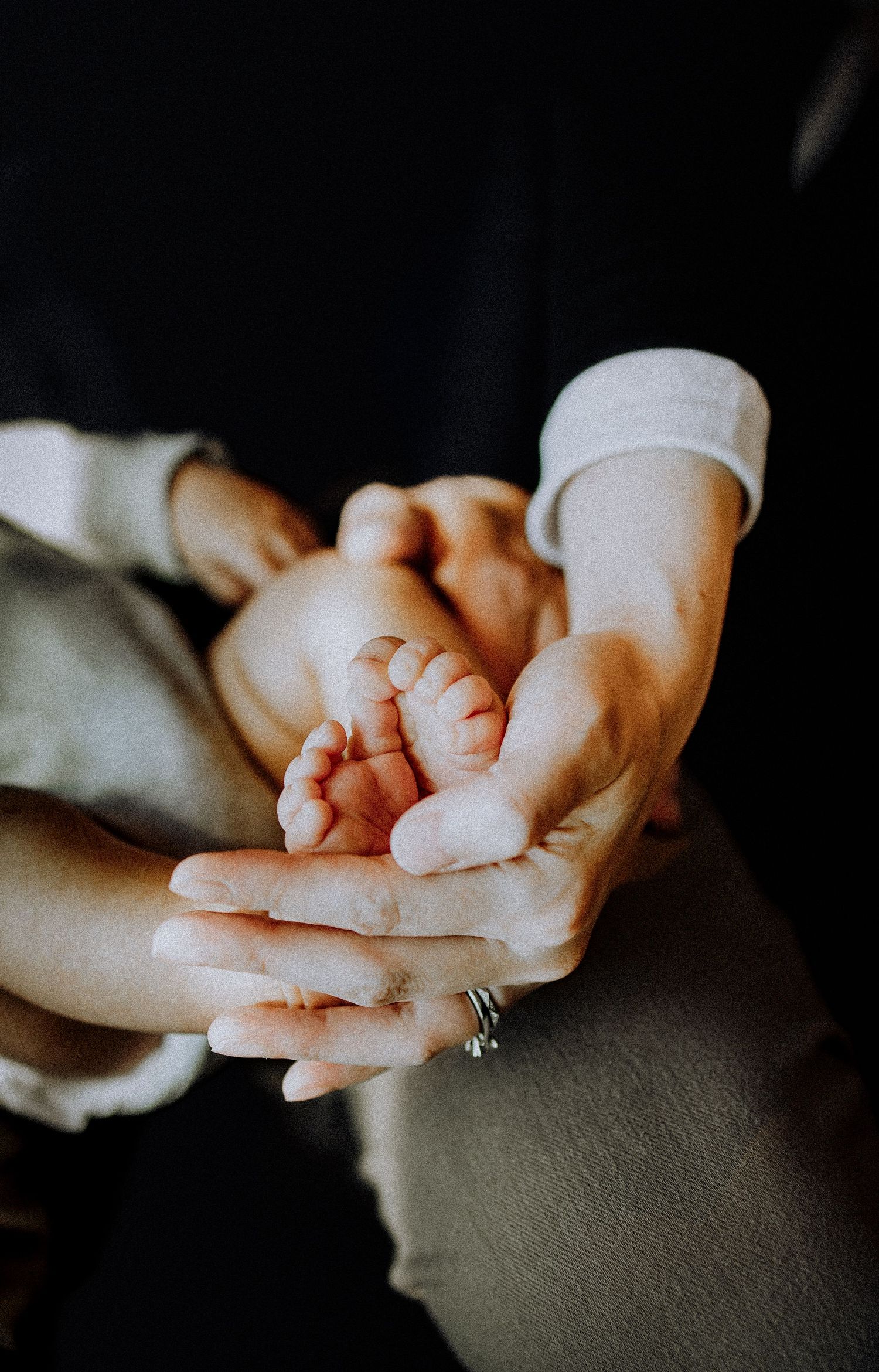 New Jersey Family Photographer-A pair of hands gently cradle tiny baby feet against a dark background, with a wedding ring visible.