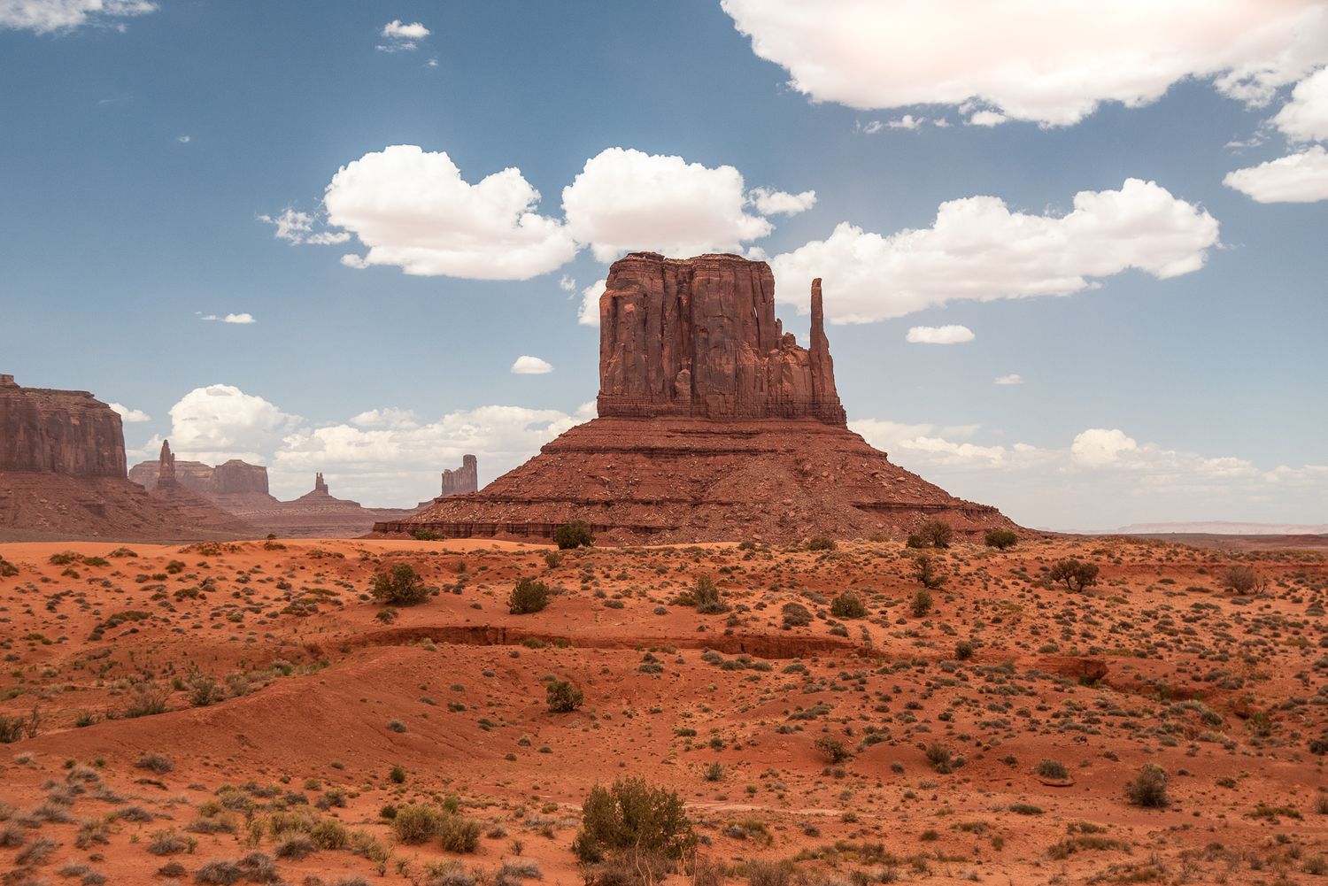 A landscape photograph of Majestic sandstone butte rises from desert landscape in Monument Valley under blue sky with white clouds.
