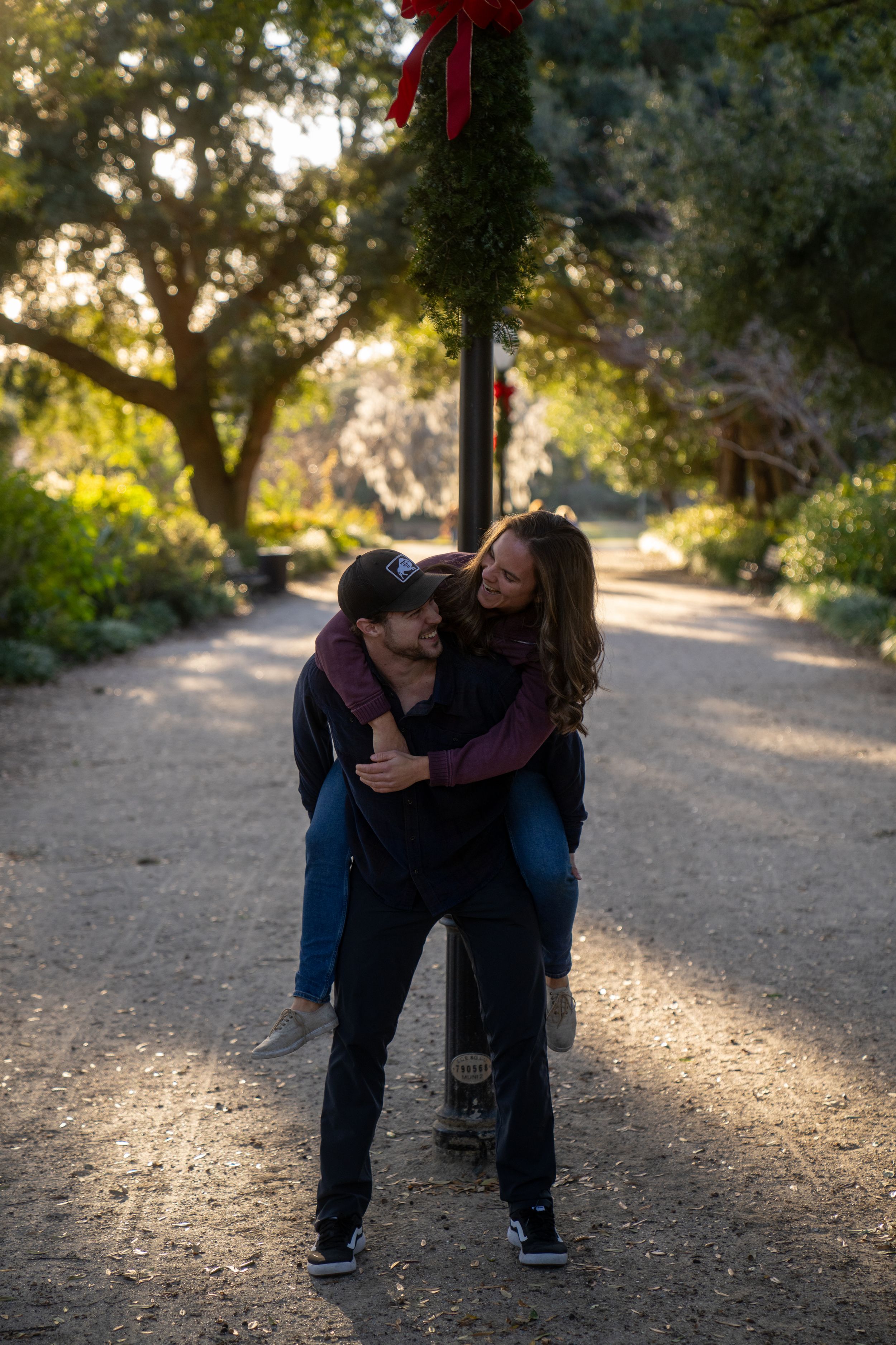 A playful piggyback ride moment captured on a tree-lined path with a festive red bow decoration.