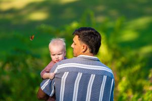 Parent lifting happy baby in air against green summer background.