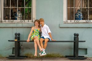 Two young children sit together on a black bench outside a building with decorative windows.