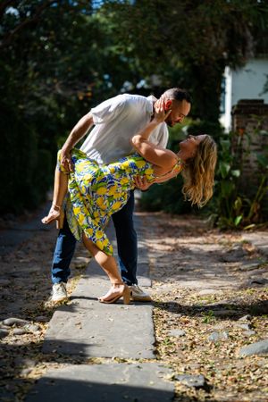 A romantic couple in casual attire share a dance move on a leaf-covered sidewalk.