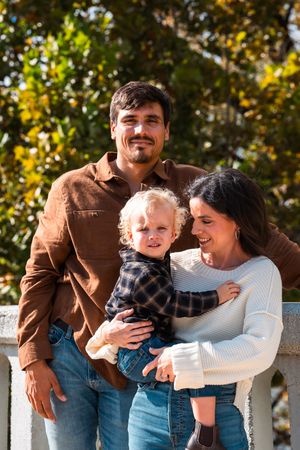 Happy young family with toddler posing outdoors in fall, parents smiling in casual attire with autumn foliage background.