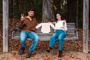 Couple sitting on wooden swing holding baby outfit, making peace signs, surrounded by autumn leaves.