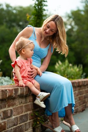 Woman in blue dress and child sit together on brick wall in garden setting.