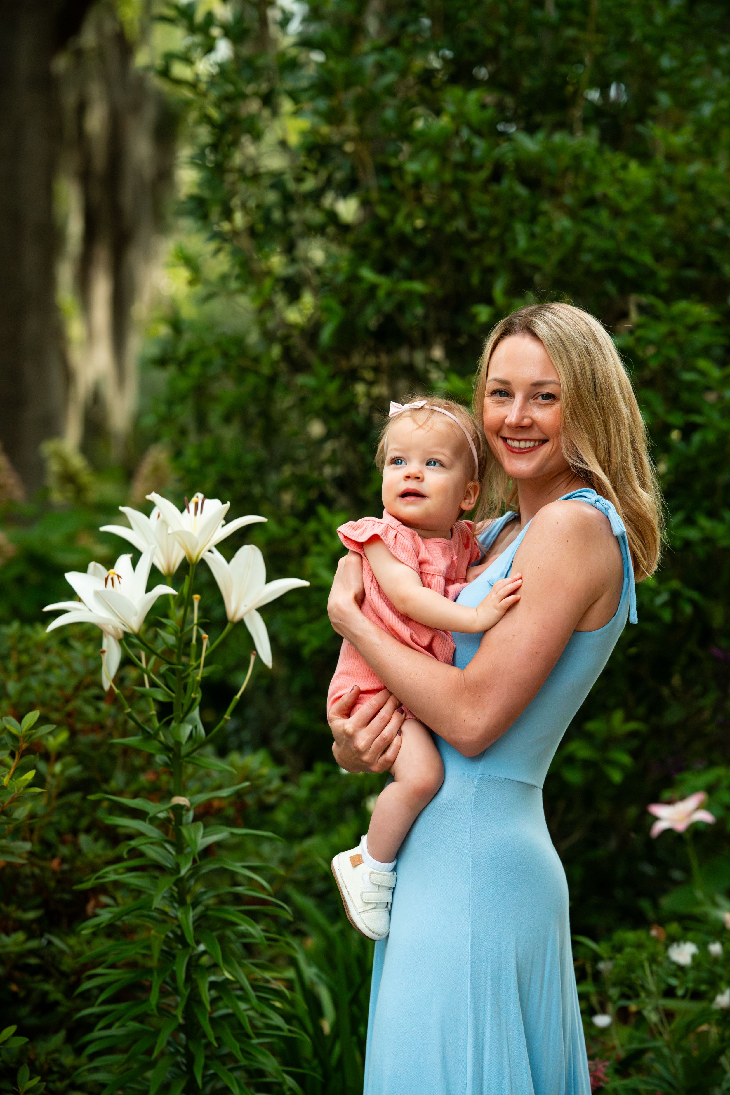 Mother and child share tender moment near blooming white lilies in garden.