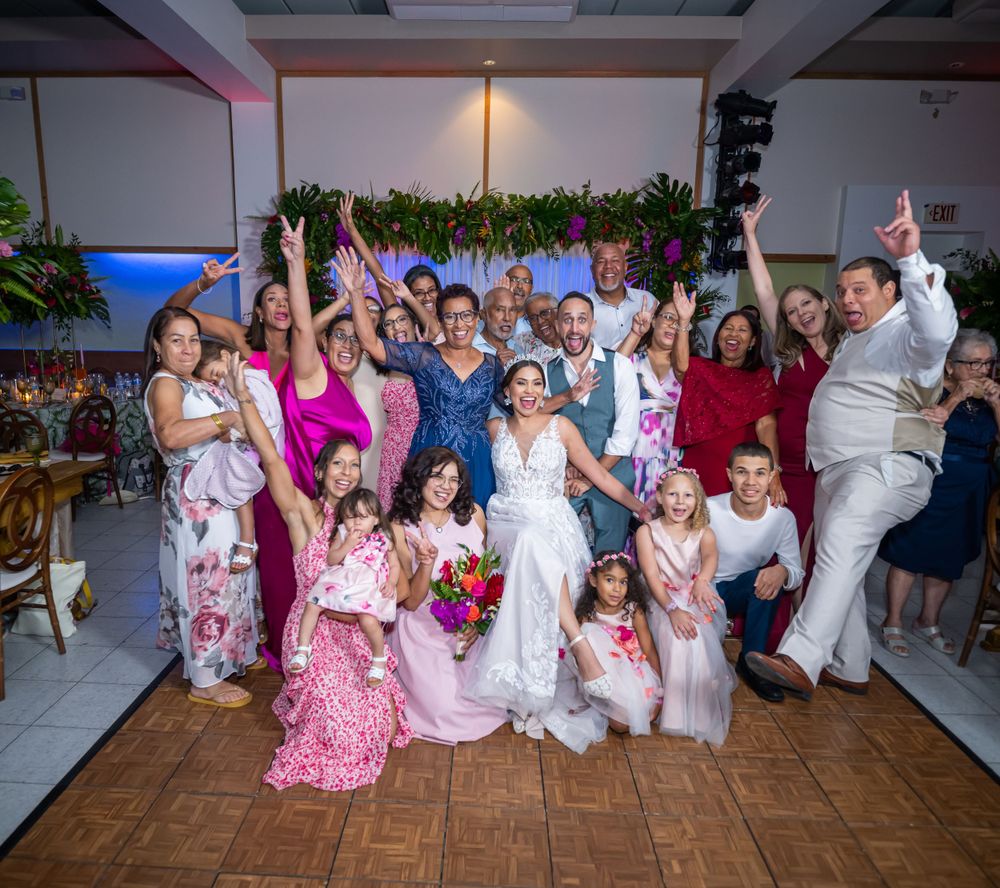 A large group wedding photo with guests in pastel attire celebrating at an indoor reception.