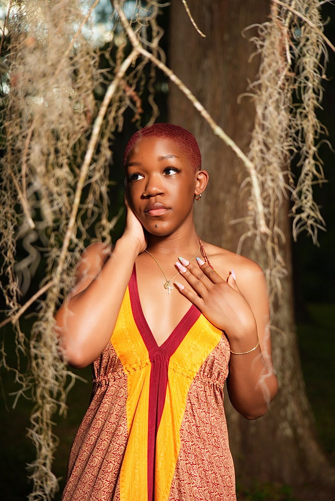 A young woman with short dyed red hair poses outdoors in an orange and yellow dress beside hanging Spanish moss.