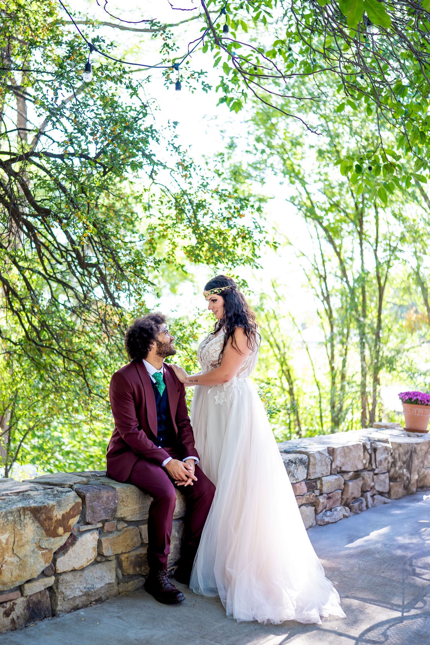 A romantic wedding photo captures a couple on a stone wall path surrounded by lush greenery.