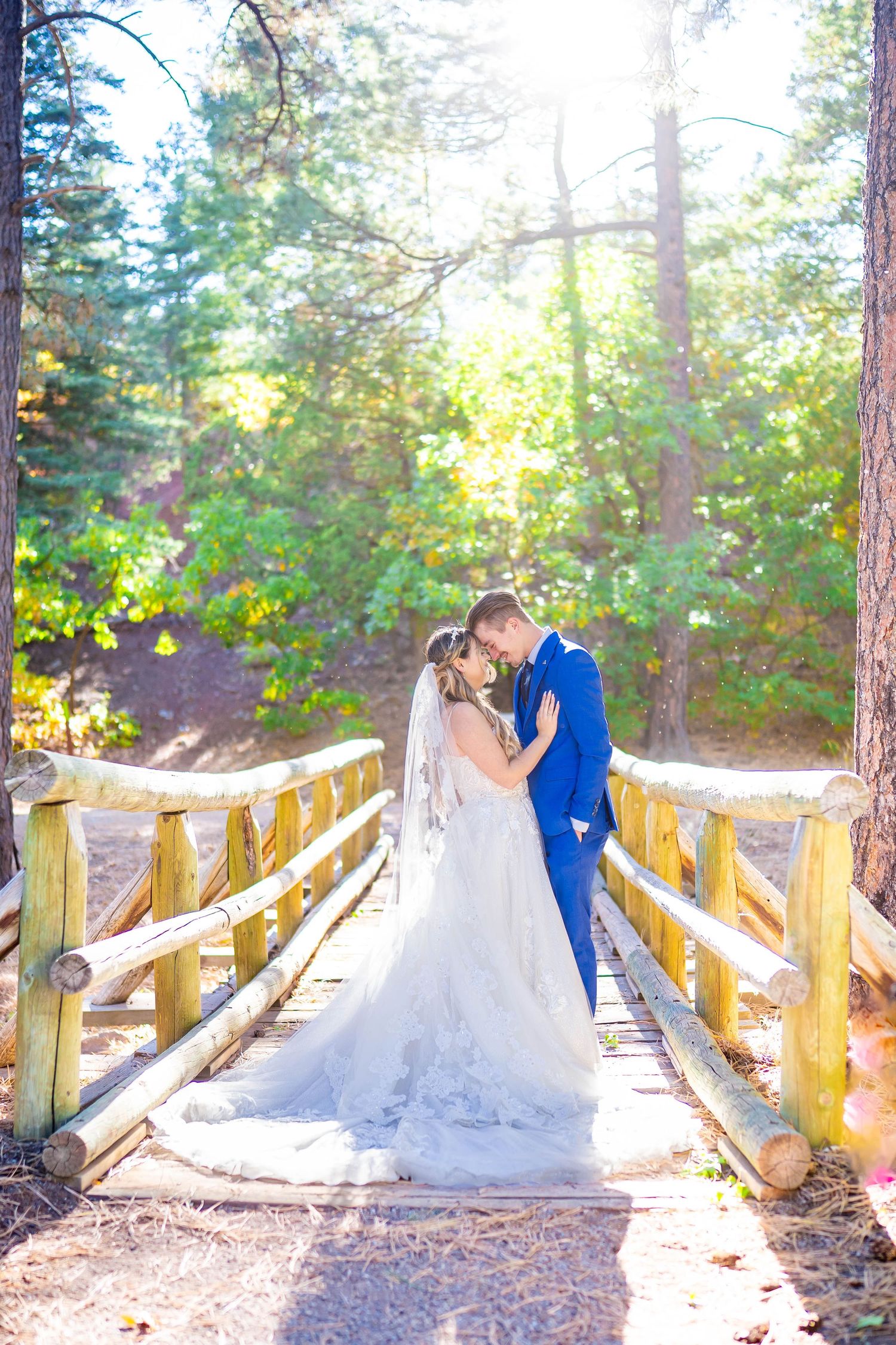 Newlyweds embrace on wooden bridge surrounded by sunlit forest.