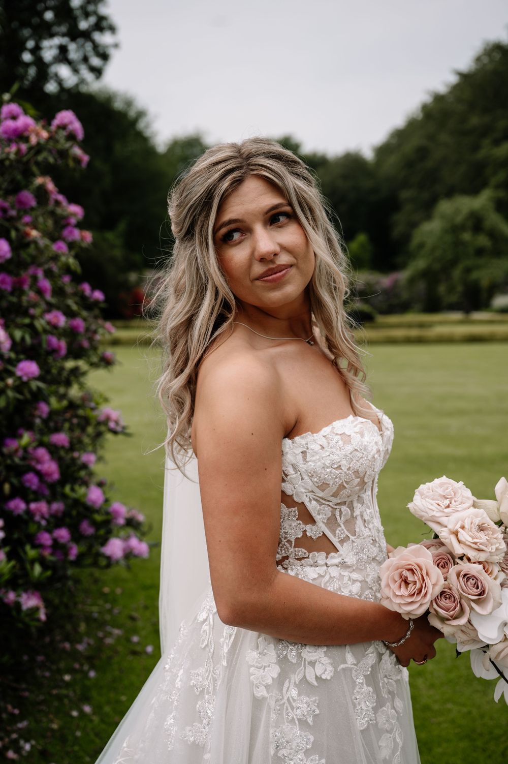Bride in lace dress poses with pink roses near flowering garden.