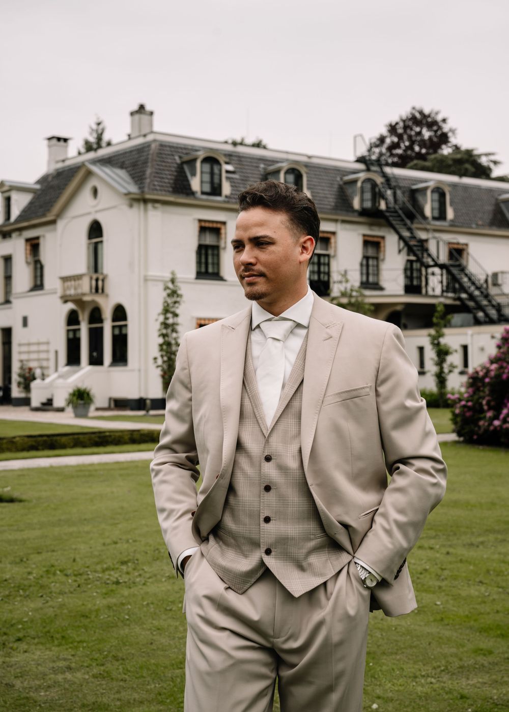 An elegant groom in a light beige three-piece suit poses in front of a grand estate mansion.