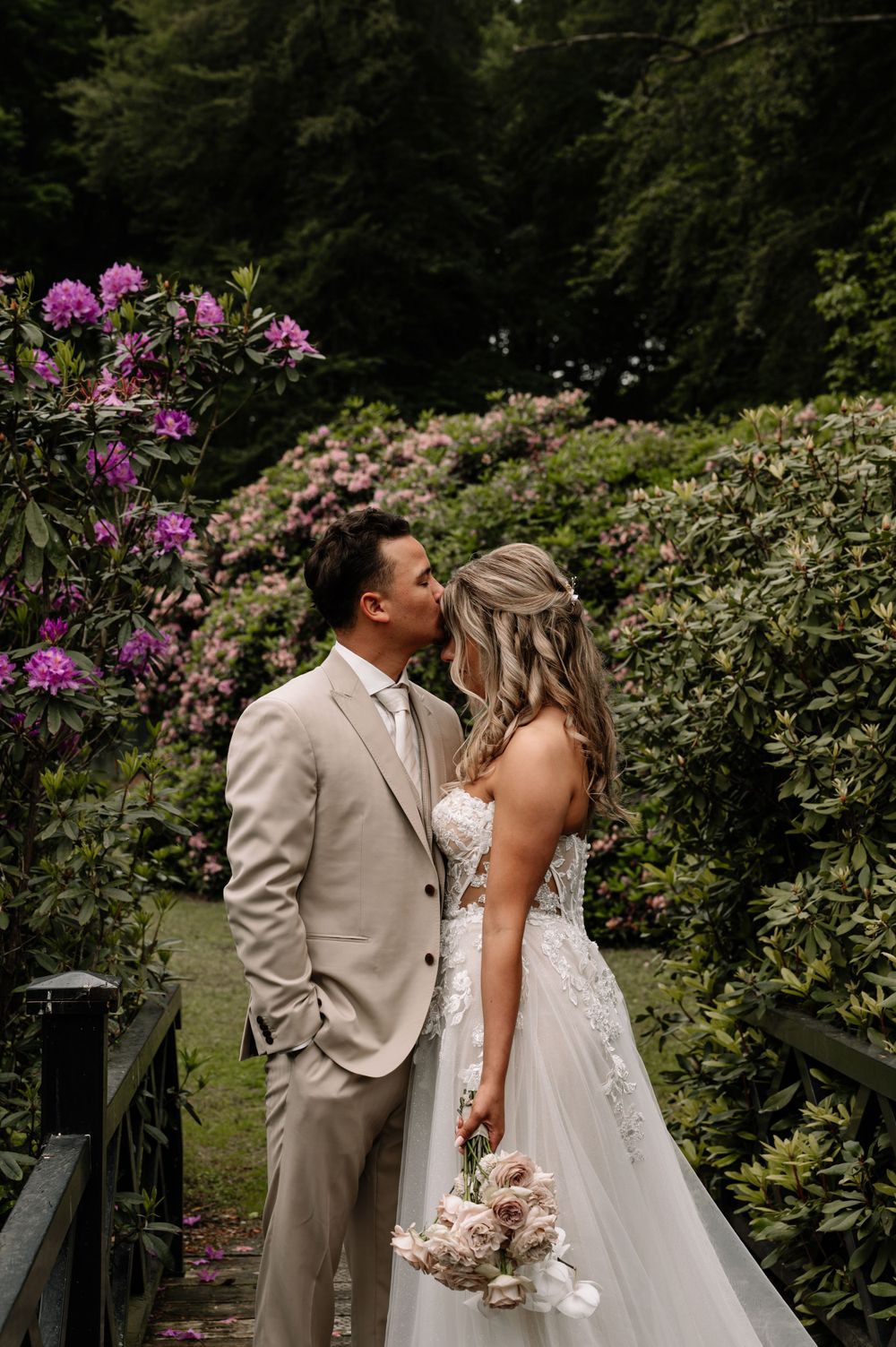 Bride and groom stand together on garden path surrounded by pink rhododendrons.