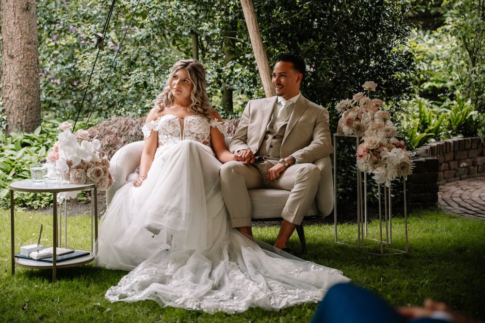 A couple sits together on outdoor furniture surrounded by elegant floral arrangements at their garden wedding.
