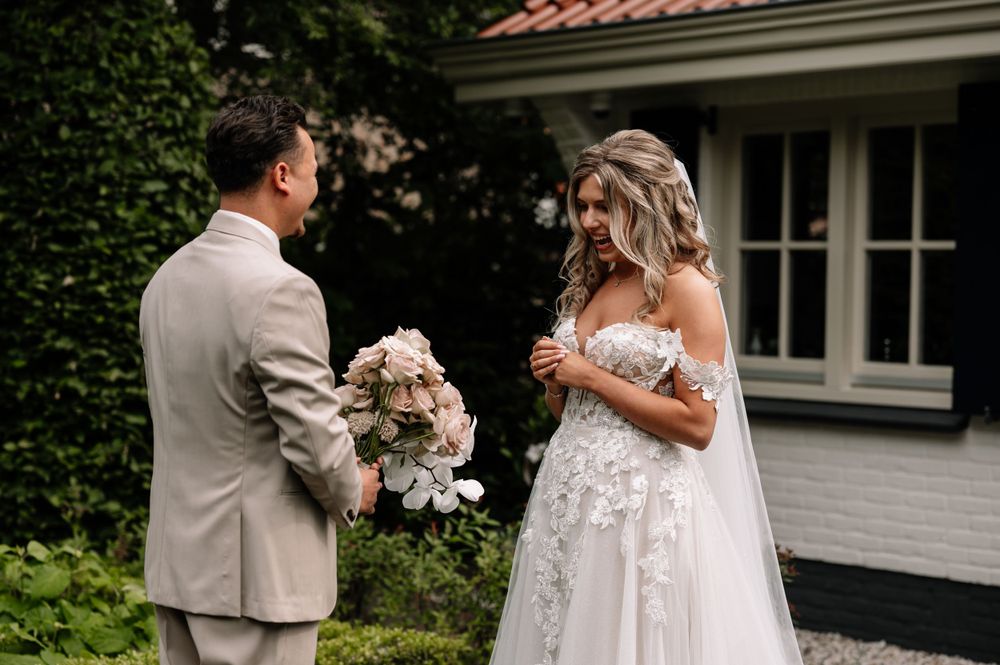 A couple shares a first look moment in front of a white house surrounded by greenery.