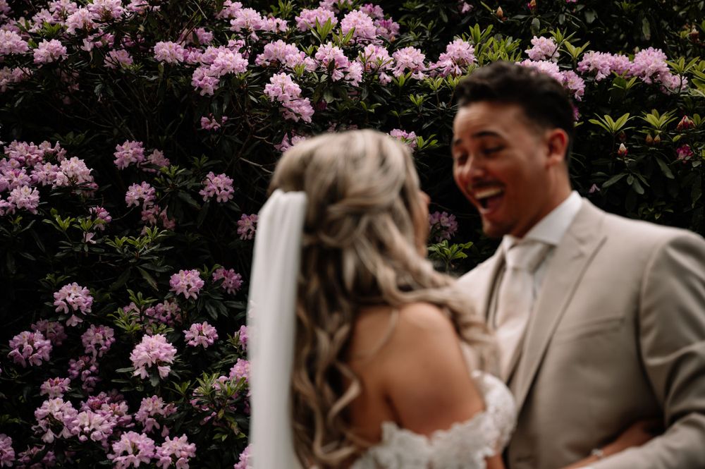 Joyful wedding couple sharing a laugh against pink flowering bushes.
