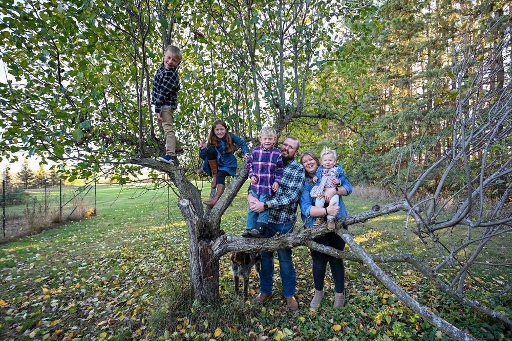 Family of six poses around a gnarled apple tree in a grassy yard, with children climbing branches and parents holding a baby in autumn.