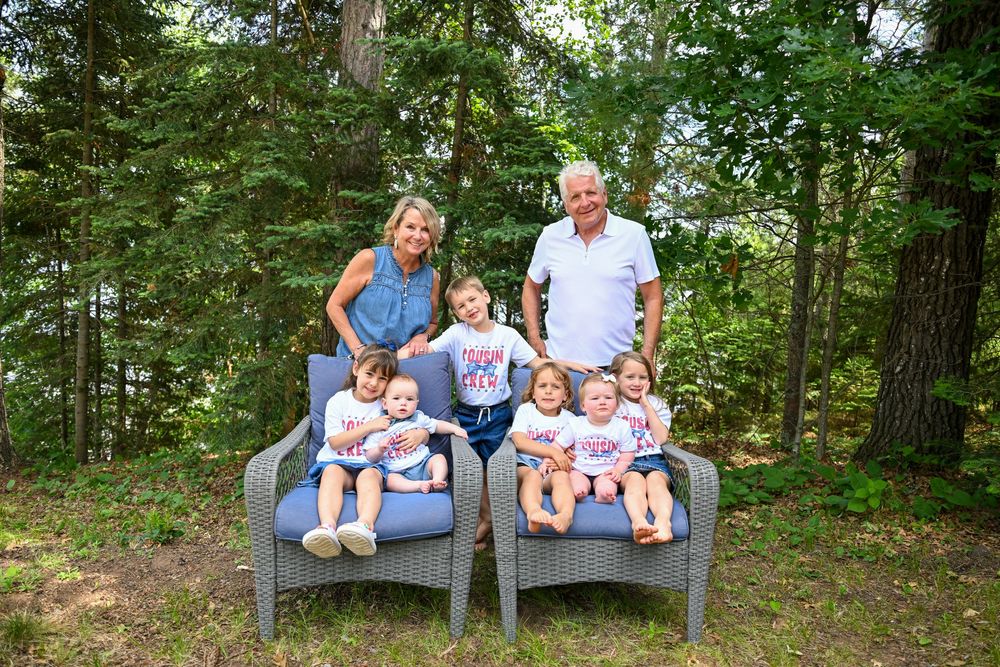 Grandparents pose with five young grandchildren in matching shirts on outdoor chairs surrounded by lush green trees.