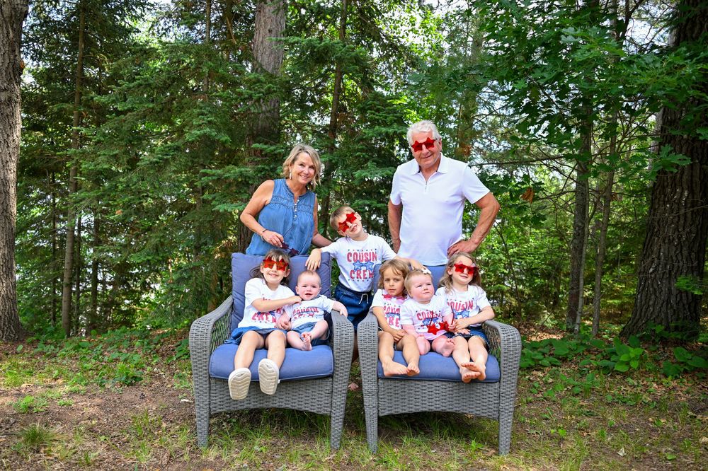Family of two adults and four young girls in matching outfits and red sunglasses posing outdoors among tall green trees.