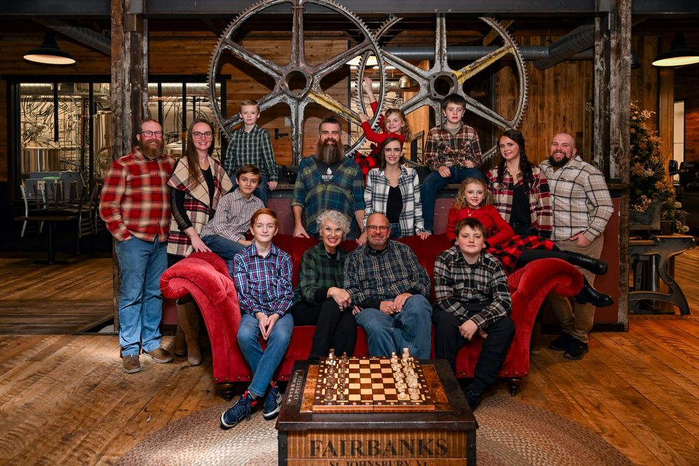 Large family group in plaid shirts posing around a Fairbanks chess table in a rustic lodge setting.