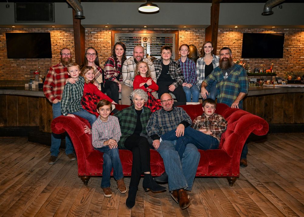 Extended family of fifteen posing together on and around a red velvet sofa in a warm restaurant setting.