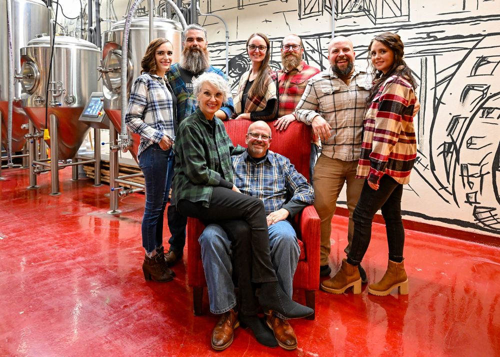 Group of seven adults posing together in front of shiny brewery tanks on a bright red floor.